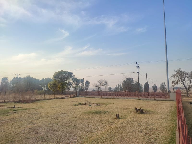 A view of a vacant residential plot featuring dry grass, perimeter fencing, and utility infrastructure under a clear sky.