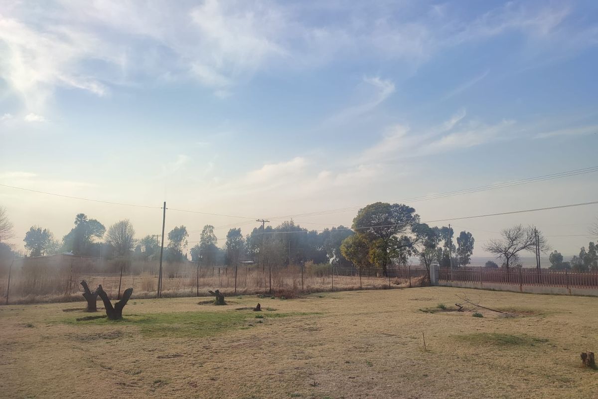A view of a vacant residential plot featuring open grass, perimeter fencing, and mature trees under a clear sky.