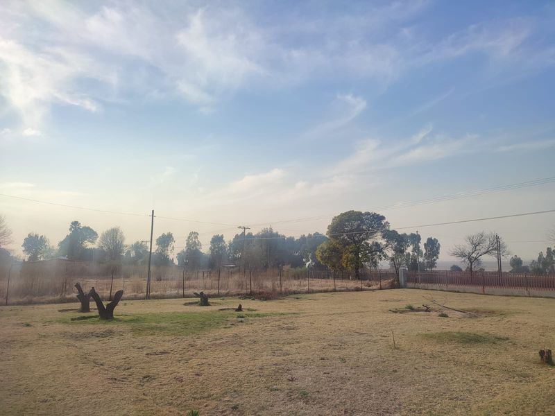 A view of a vacant residential plot featuring open grass, perimeter fencing, and mature trees under a clear sky.
