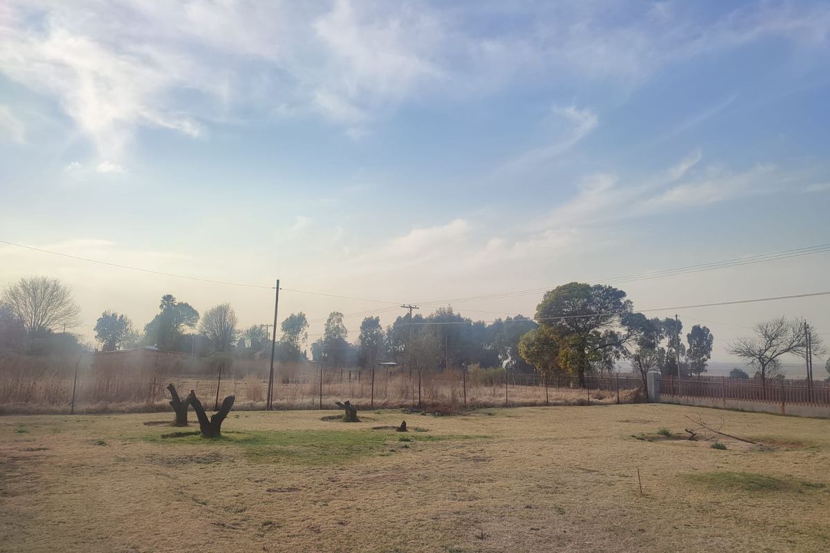 A view of a vacant residential plot featuring dry grass, wire mesh perimeter fencing, and scattered trees under a clear sky.