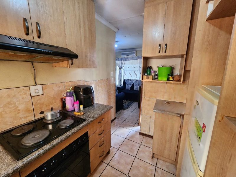 Compact kitchen featuring built-in cabinetry, electric hob, and extractor fan, with tiled flooring and splashbacks, leading into an adjacent living area.