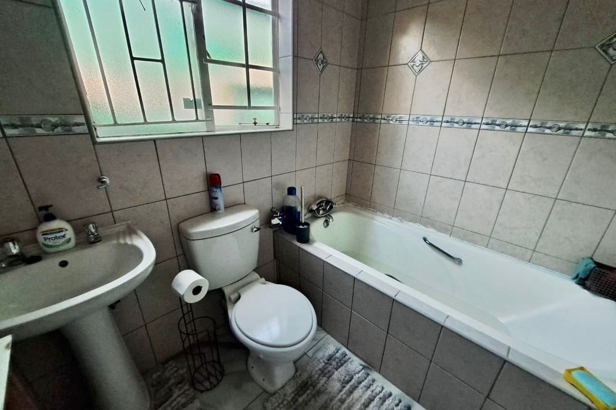 A functional bathroom featuring a pedestal basin, toilet, and built-in bathtub with tiled walls and a window with security bars.