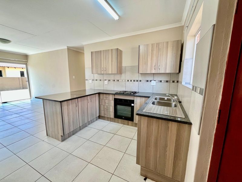 A clean, functional kitchen featuring wood-look cabinetry, dark countertops, a built-in oven, gas hob, and tiled flooring, flowing into an open-plan living area.