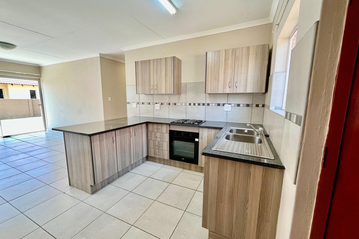 A clean, functional kitchen featuring wood-look cabinetry, dark countertops, a built-in oven, gas hob, and tiled flooring, flowing into an open-plan living area.
