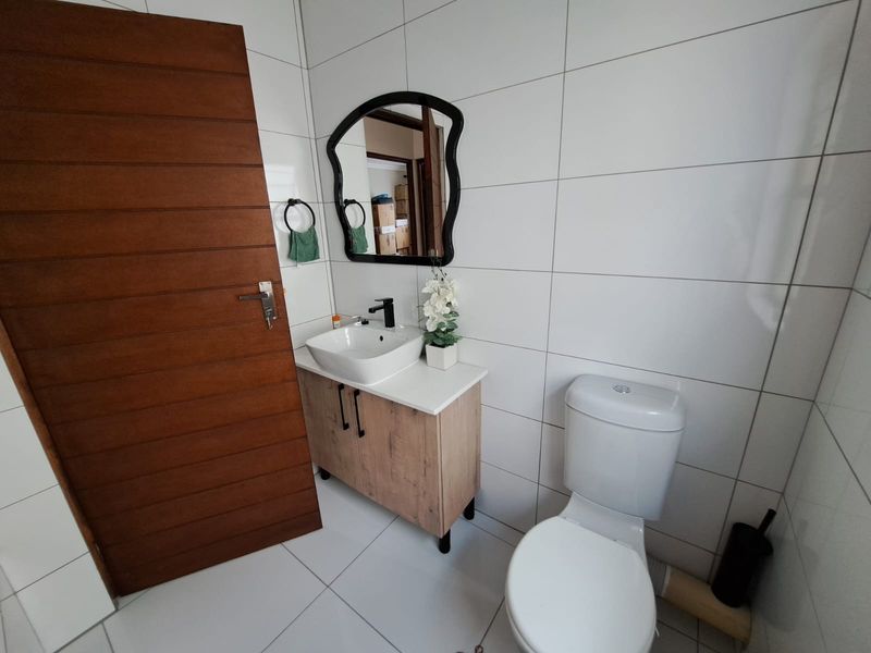 A modern bathroom featuring a wood-finish vanity cabinet, vessel sink, wall-mounted mirror, and white tiled walls and flooring.