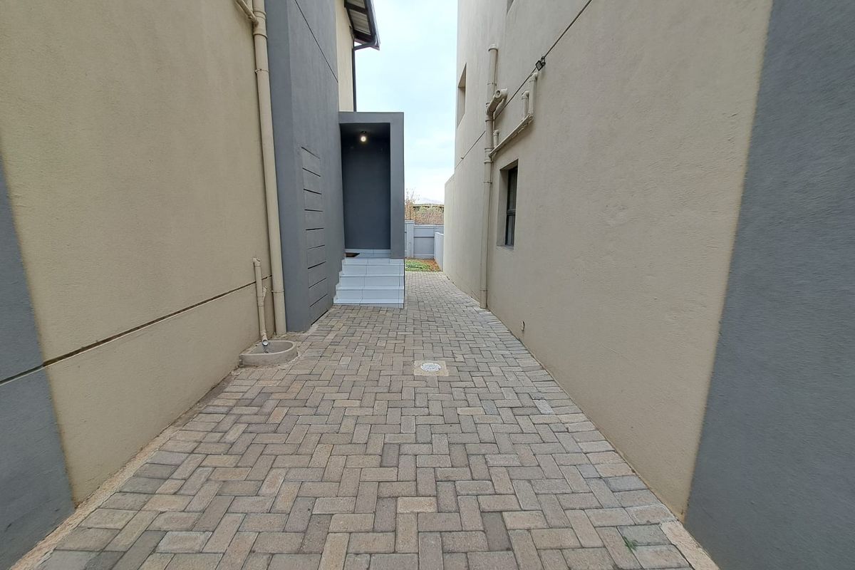 A paved side walkway between two townhouse structures, featuring external plumbing, tiled steps leading to an entrance, and modern exterior wall finishes.