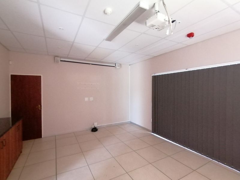 A functional cinema room featuring a suspended ceiling, mounted projector, projection screen, tiled flooring, and vertical blinds.