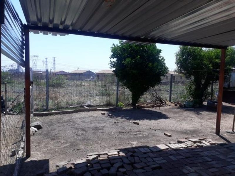 A covered outdoor patio area featuring a corrugated metal roof, metal support posts, and basic brick paving overlooking an unlandscaped yard.