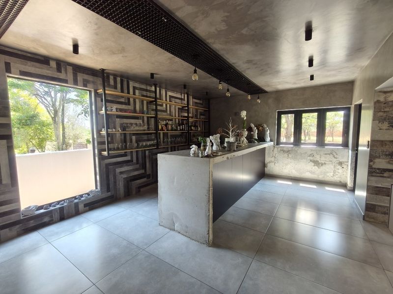 A modern bar area featuring a concrete counter, industrial-style shelving, decorative geometric wall tiles, and large windows providing natural light.