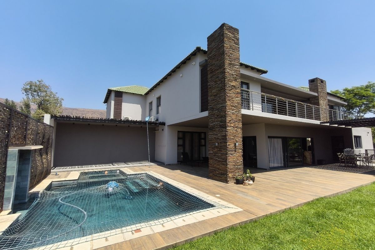 Rear elevation of a modern double-story house featuring a swimming pool, expansive timber decking, stone-clad chimneys, and a spacious upper-level balcony.