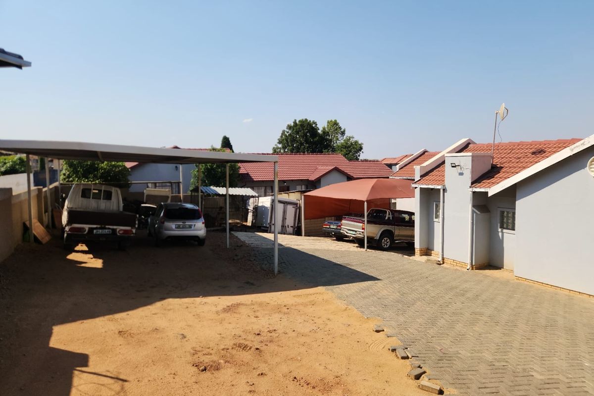 Rear exterior view featuring a paved driveway, covered carport parking, and tiled roof structure with adjacent sandy ground area.