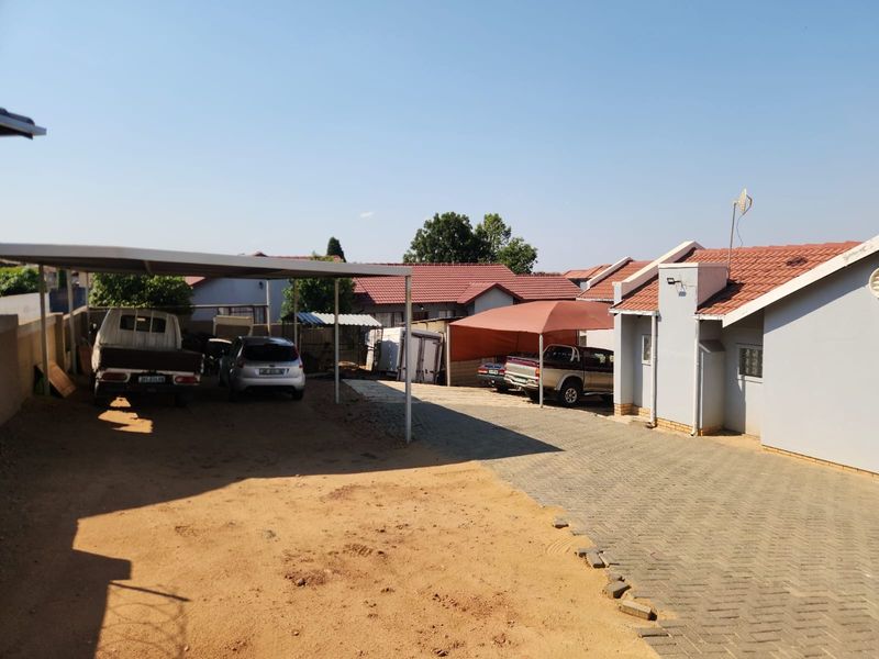 Rear exterior view featuring a paved driveway, covered carport parking, and tiled roof structure with adjacent sandy ground area.