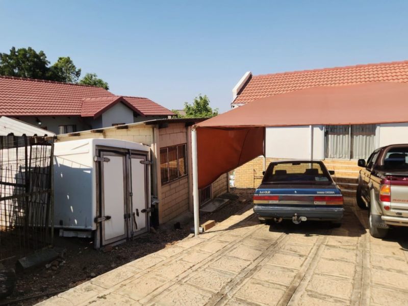 Rear exterior view showing a paved driveway, a shade cloth carport structure, and a small brick outbuilding under a tiled roof.