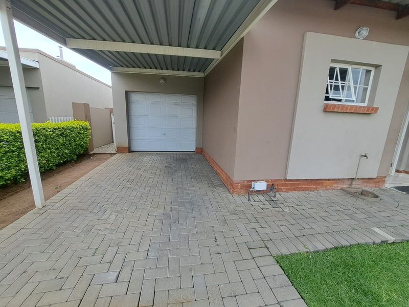 A paved driveway leading to a single garage with an attached metal carport, featuring a neat brick facade and exterior water connection.