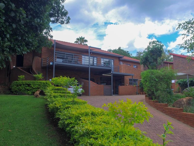 A multi-level brick house featuring a raised balcony, paved driveway, and established garden landscaping.