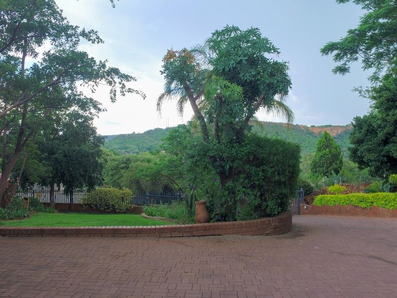 A well-maintained garden area featuring a brick-paved driveway, low brick retaining walls, and established trees with scenic hill views in the background.