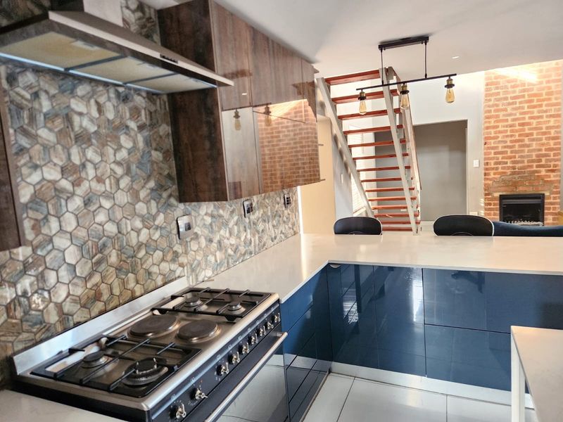 Modern kitchen featuring high-gloss cabinetry, stone countertops, and a mosaic backsplash, seamlessly flowing into an open-plan living area with a feature brick fireplace.