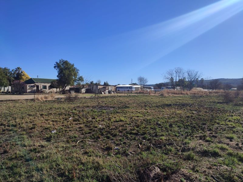 A view of an undeveloped residential plot of land with surrounding fencing and nearby structures visible in the distance.