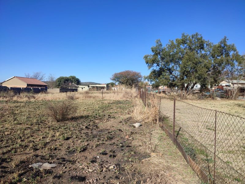 A view of a vacant residential plot featuring wire mesh fencing and mature trees, showing natural ground cover and surrounding neighborhood structures.