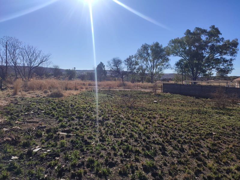 A view of a vacant residential plot featuring natural ground cover, perimeter fencing, and a retaining wall under a clear sky.