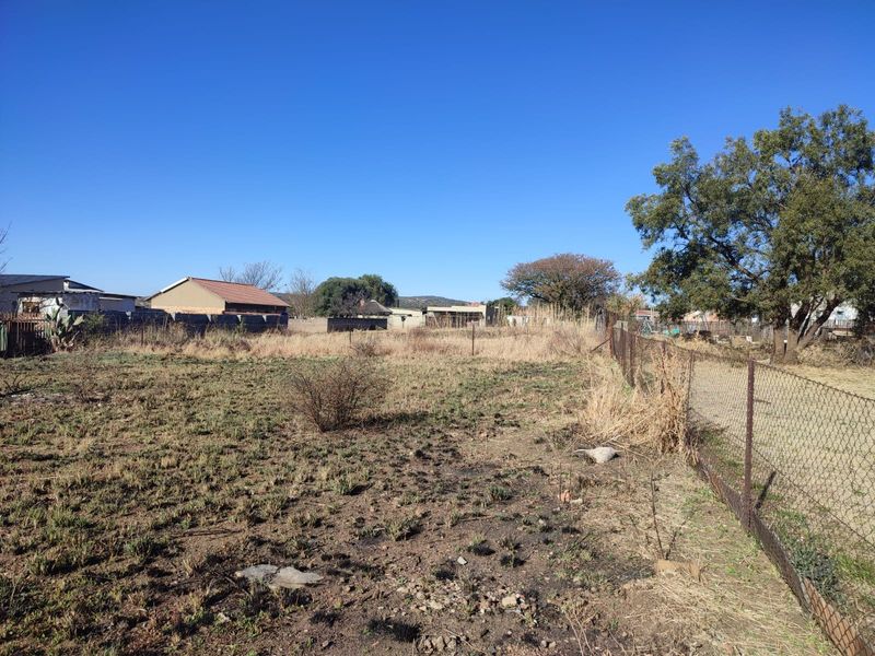 A view of a vacant residential plot featuring a chain-link boundary fence and surrounding residential properties in the background.