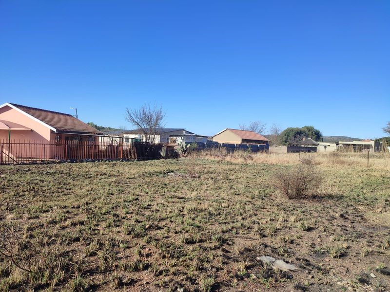 A view of a vacant residential plot featuring dry grass and surrounding neighboring properties with metal fencing.