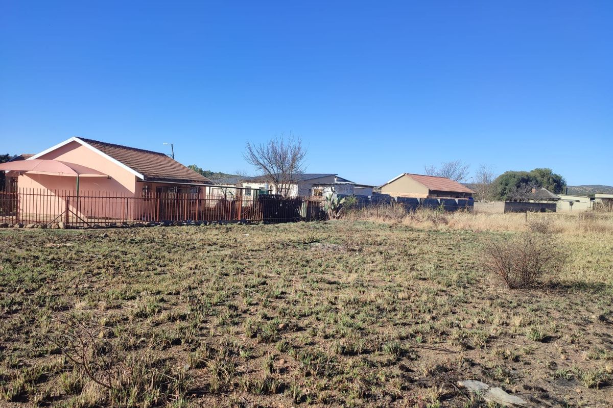 A view of the rear exterior of the residential property, featuring a large vacant plot, metal perimeter fencing, and existing dwelling structures.