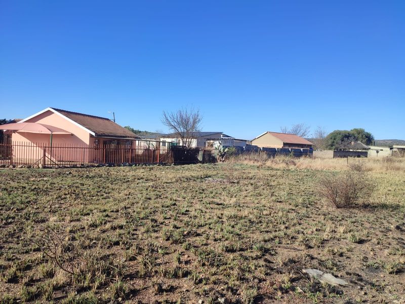 A view of the rear exterior of the residential property, featuring a large vacant plot, metal perimeter fencing, and existing dwelling structures.