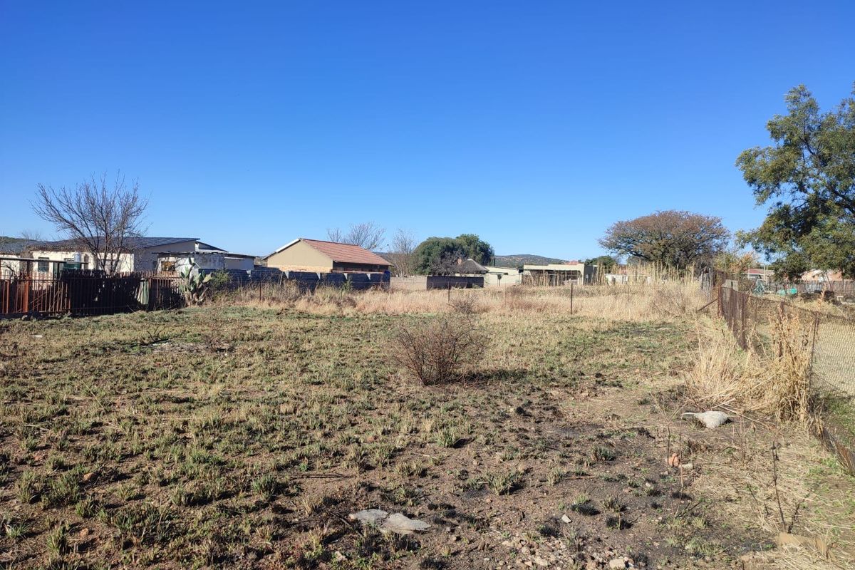 A vacant residential plot featuring dry, overgrown grass, enclosed by wire mesh fencing and a metal gate, situated in a suburban neighborhood.