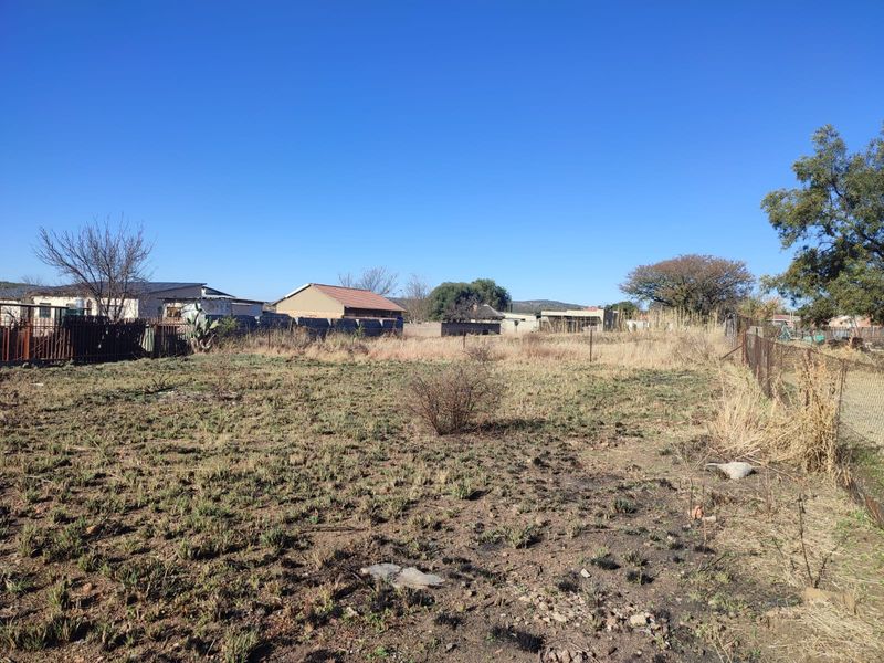 A vacant residential plot featuring dry, overgrown grass, enclosed by wire mesh fencing and a metal gate, situated in a suburban neighborhood.