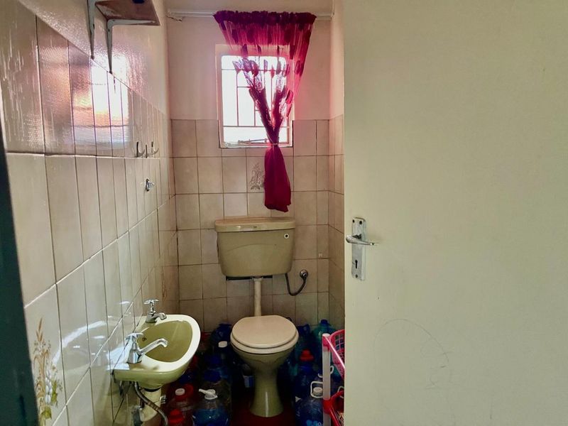 A compact bathroom featuring dated ceramic sanitaryware and wall tiling, with a small window fitted with security bars.