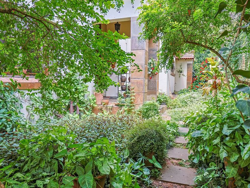 A lush, well-established garden featuring a stone-paved pathway leading to a covered patio entrance with stone-clad pillars.