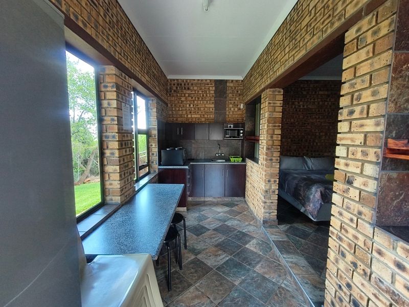 A compact kitchen area featuring exposed brick walls, tiled flooring, built-in cabinetry, and a stone-look countertop, adjacent to a bedroom space.