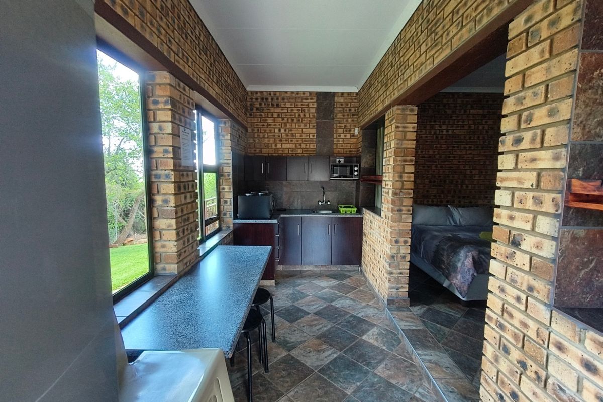 A compact kitchen area featuring exposed brick walls, tiled flooring, built-in cabinetry, and a stone-look countertop, adjacent to a bedroom space.