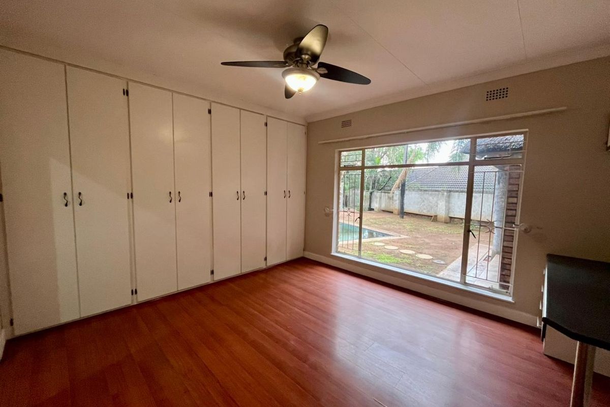 Spacious bedroom featuring extensive built-in cupboards, laminate flooring, a ceiling fan, and a large window overlooking the garden.
