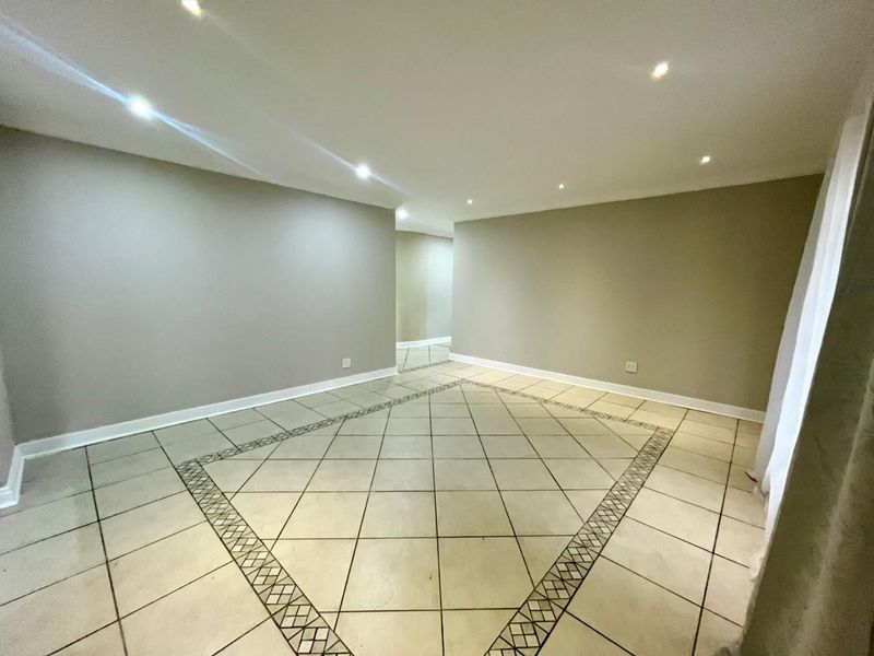 A spacious, empty living area featuring neutral wall tones, recessed downlighting, and ceramic floor tiles with a decorative border.
