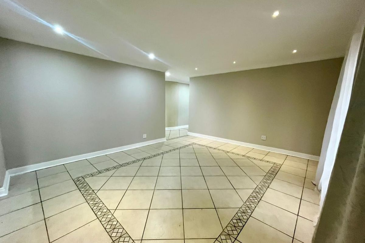 A spacious, empty living area featuring neutral wall tones, recessed downlighting, and ceramic floor tiles with a decorative border.