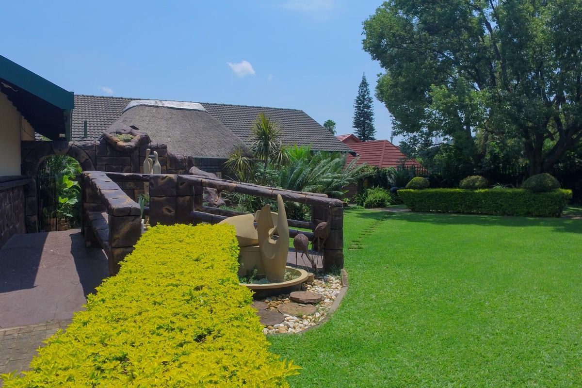A well-maintained rear garden featuring a lush lawn, mature trees, a stone-clad feature wall, and a combination of thatched and tiled roof structures.