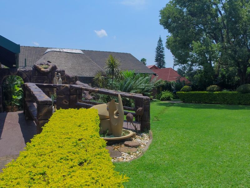 A well-maintained rear garden featuring a lush lawn, mature trees, a stone-clad feature wall, and a combination of thatched and tiled roof structures.