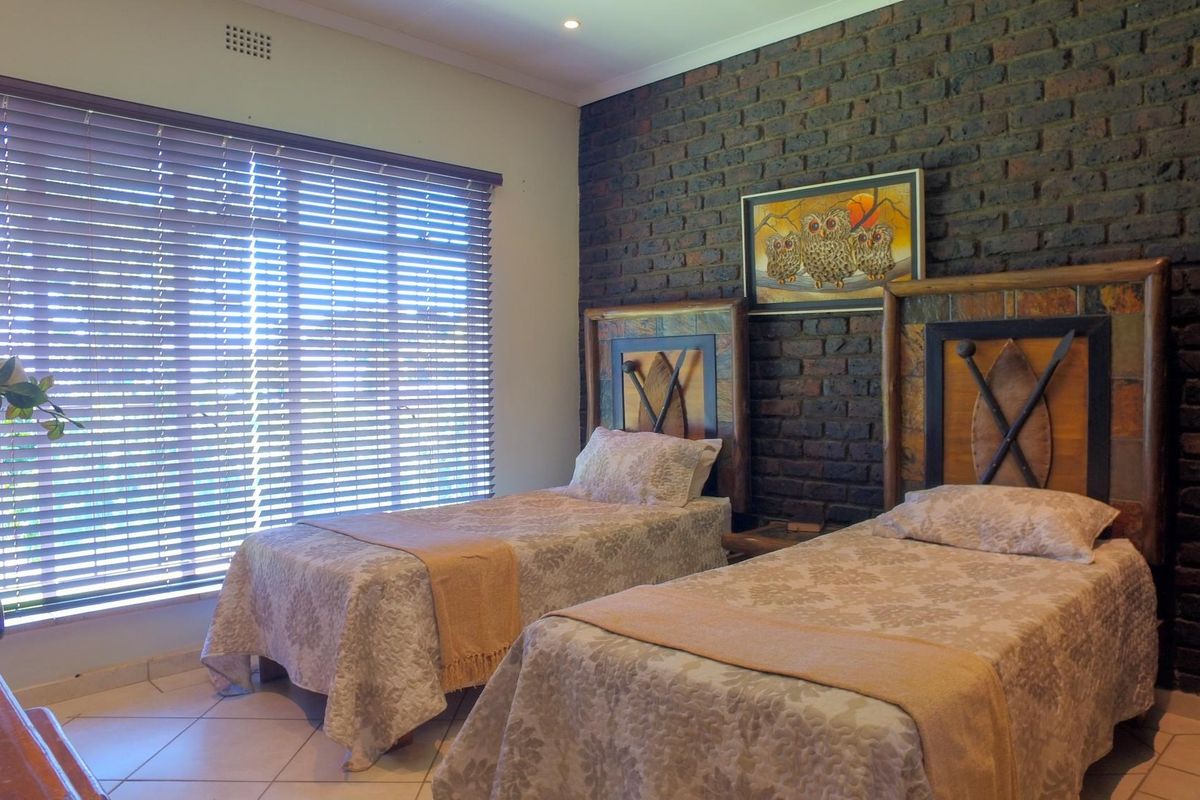 A well-lit bedroom featuring a distinctive exposed brick feature wall, tiled flooring, and large windows fitted with venetian blinds.