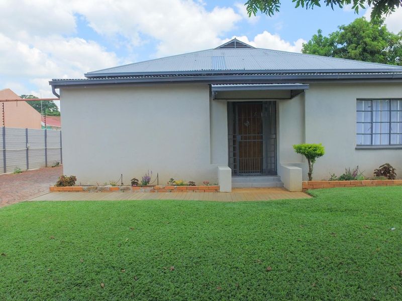 Front view of a single-story house featuring a corrugated metal roof, security entrance gate, and a well-maintained lawn with brick-edged garden beds.