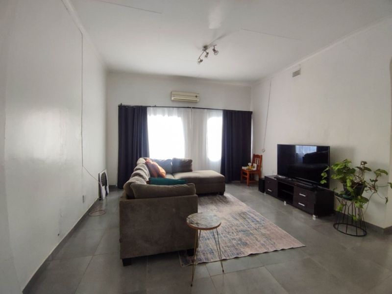 A spacious living room featuring neutral tiled flooring, a wall-mounted air conditioning unit, and ample natural light through the window.