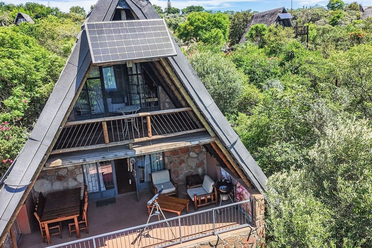 An elevated view of an A-frame house featuring stone cladding, a balcony with metal railings, and a solar panel installed on the roof.