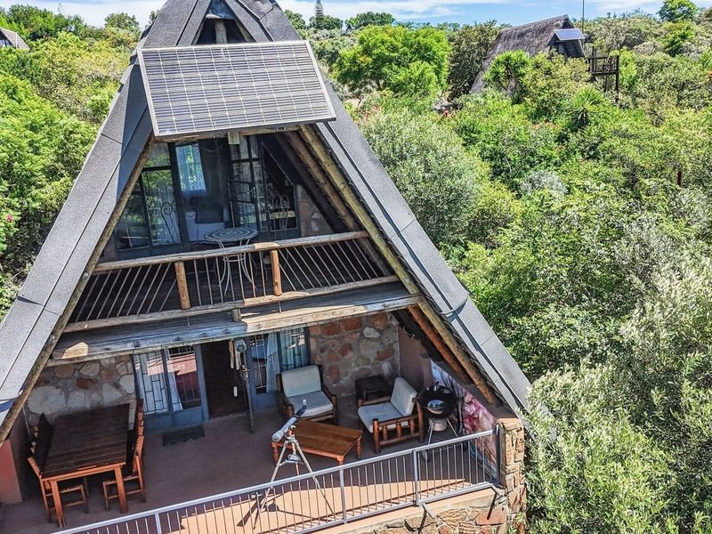 An elevated view of an A-frame house featuring stone cladding, a balcony with metal railings, and a solar panel installed on the roof.