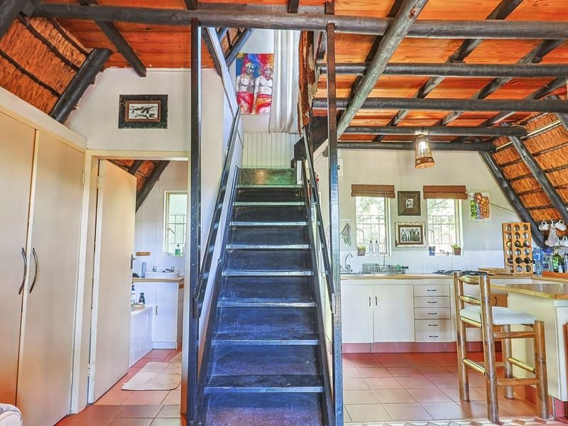 Open plan living area featuring exposed timber rafters, a thatched ceiling, tiled flooring, and a central staircase leading to an upper level.