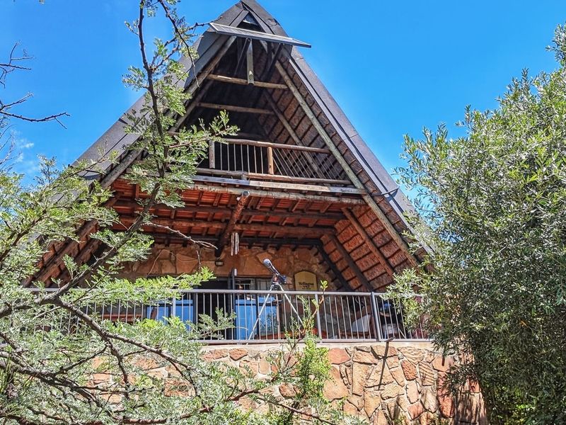 A charming A-frame house featuring rustic stone cladding, a thatched roof, and a timber balcony overlooking the surrounding natural landscape.