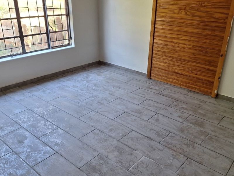 A clean, empty bedroom featuring modern grey tiled flooring, a built-in wooden cupboard, and a window fitted with security bars.