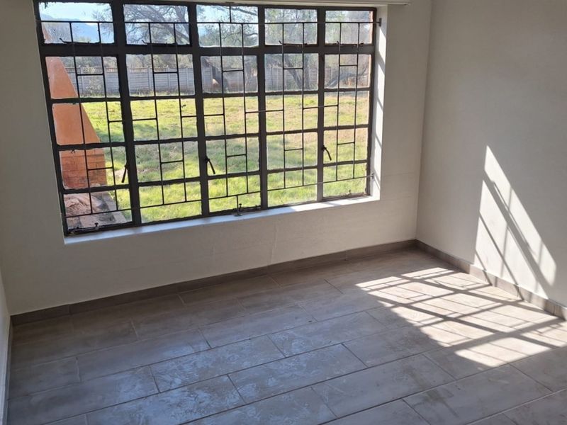 A bright, empty bedroom featuring tiled flooring and a large window with integrated steel security burglar bars, overlooking the garden.