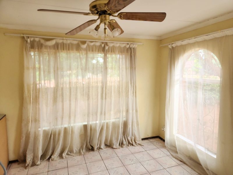 A bright, empty living area featuring ceramic tile flooring, a ceiling fan, and large windows with security bars, offering a blank canvas for renovation.