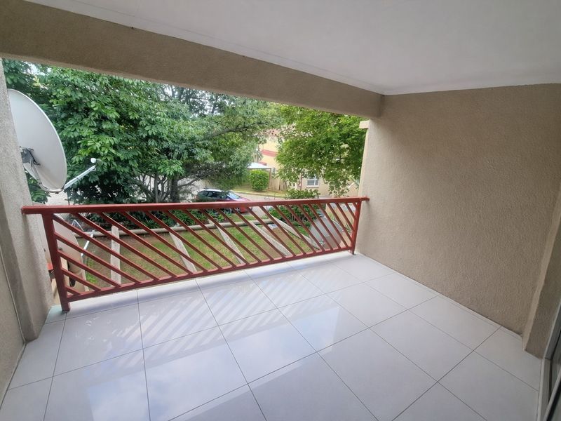 A clean, covered balcony featuring white tiled flooring and a red metal balustrade, overlooking a green garden area.